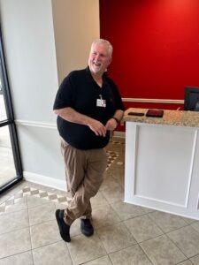 A smiling man stands in a welcoming space, wearing a black polo and ID badge, next to a reception area with a red wall.